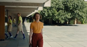 A person stands confidently in the foreground wearing a yellow shirt and red pants, while others walk in the background.