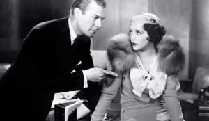 A man in a suit gestures accusatorily at a woman in a fur stole and a bow-tied blouse who looks back at him with a surprised expression in a vintage, black and white setting.