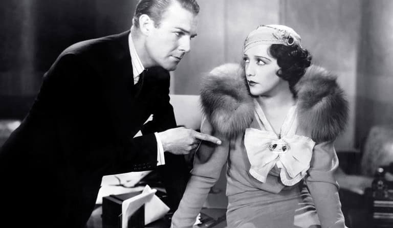 A man in a suit gestures accusatorily at a woman in a fur stole and a bow-tied blouse who looks back at him with a surprised expression in a vintage, black and white setting.