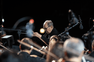 ‘Ennio’ Blu-Ray Review – Cinematic Icon Gets His Time In The Spotlight A man in glasses is playing an instrument in front of a group of people.