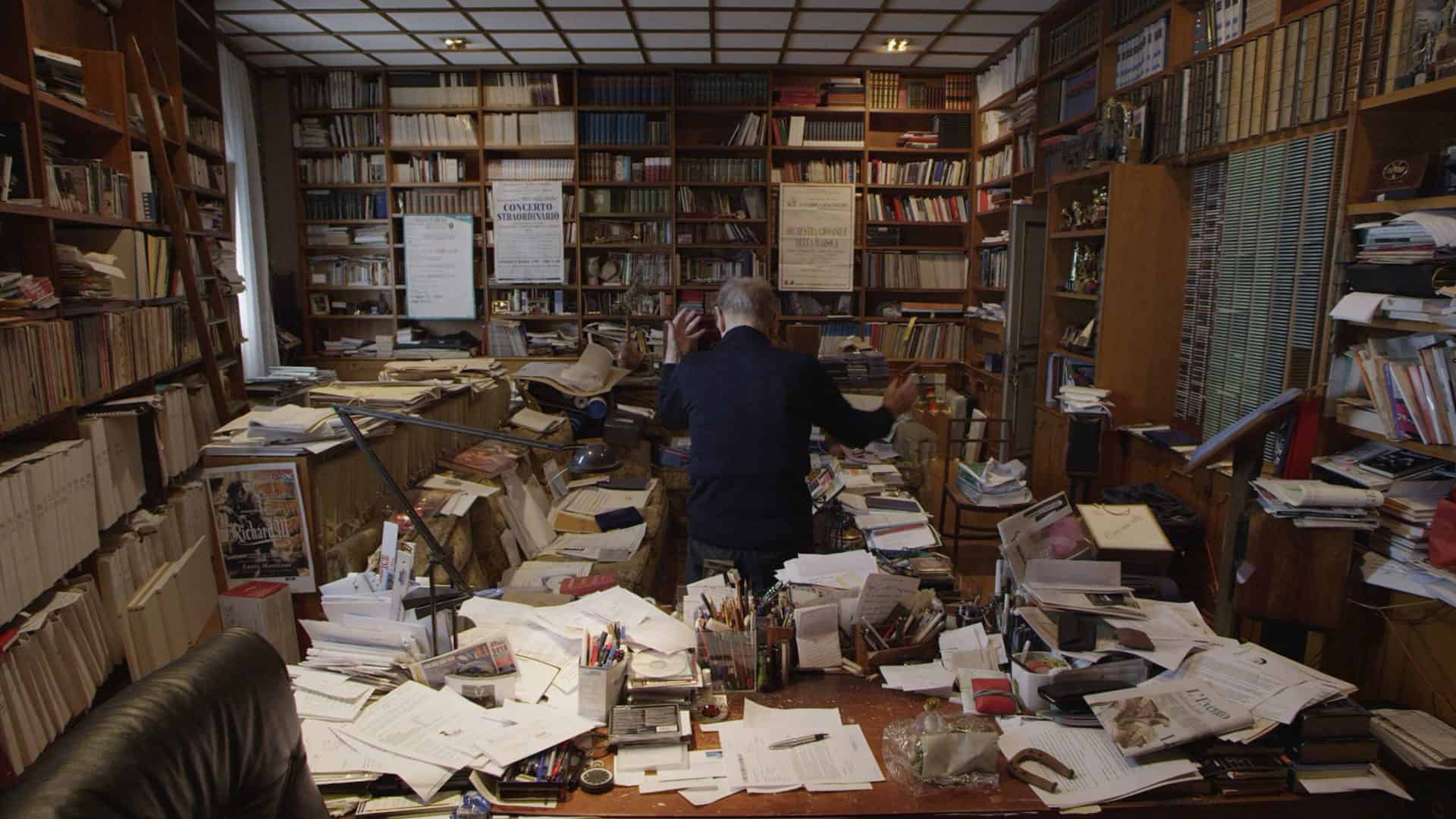A man in a suit standing in a room full of books.