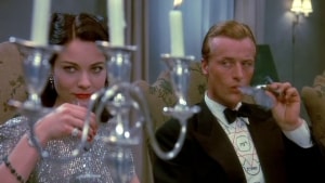 A man in a tuxedo and a woman in a shimmering dress sitting at a table with candles, both looking pensive, with the man smoking and the woman holding a cocktail.