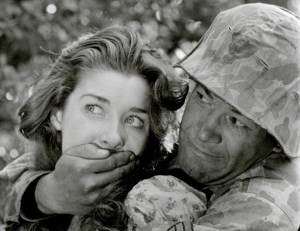 A soldier in camouflage attire gently placing his hand over a young woman's mouth.