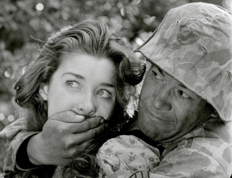 A soldier in camouflage attire gently placing his hand over a young woman's mouth.
