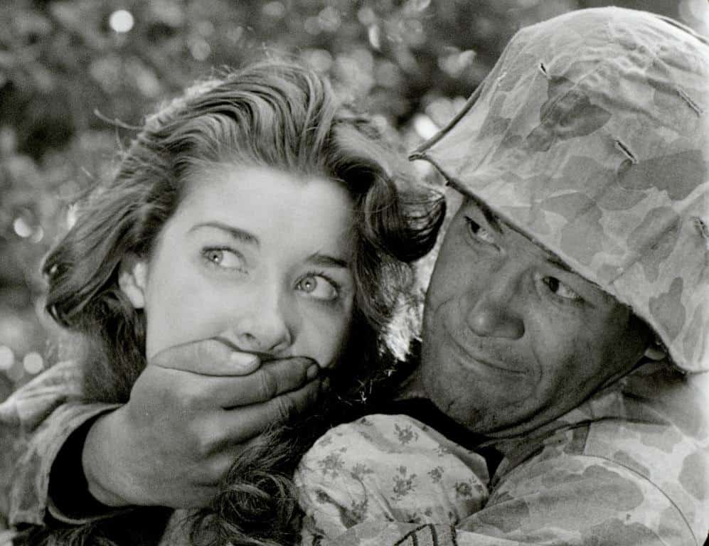 A soldier in camouflage attire gently placing his hand over a young woman's mouth.