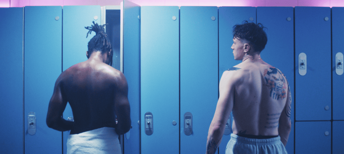 Two individuals in a locker room facing away from the camera; one is wrapped in a towel, and the other is shirtless revealing tattooed back.