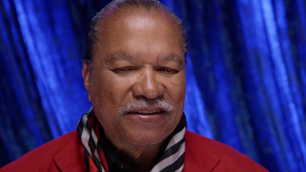 A close-up of a smiling man with a mustache wearing a red jacket against a blue draped background.
