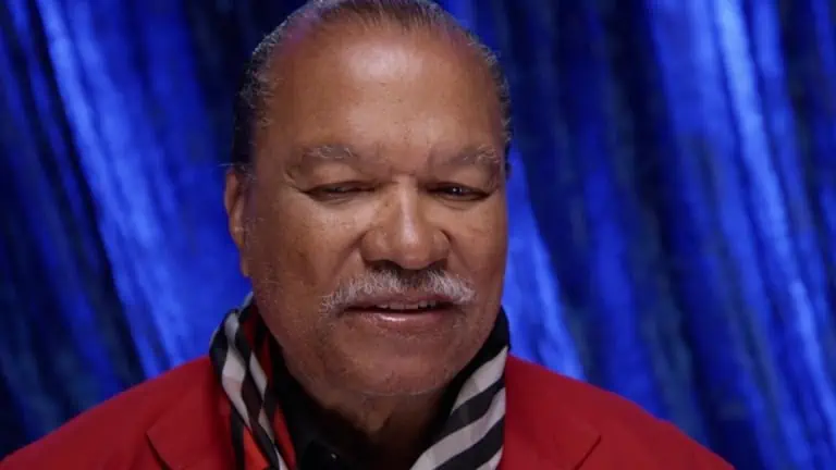 A close-up of a smiling man with a mustache wearing a red jacket against a blue draped background.