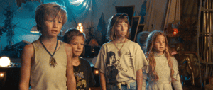 ‘Riddle Of Fire’ Blu-Ray Review – Enchanting Adventure Is One Of The Year’s True Gems Four children standing together with a mixture of emotions, such as anticipation and concern, in a dimly lit room with a vintage decor.