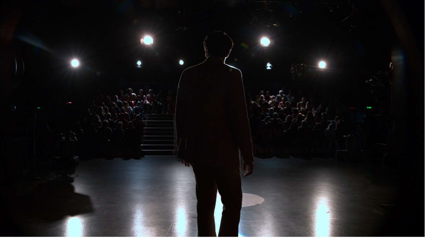 Performer facing an audience from a stage, silhouetted by bright stage lights.