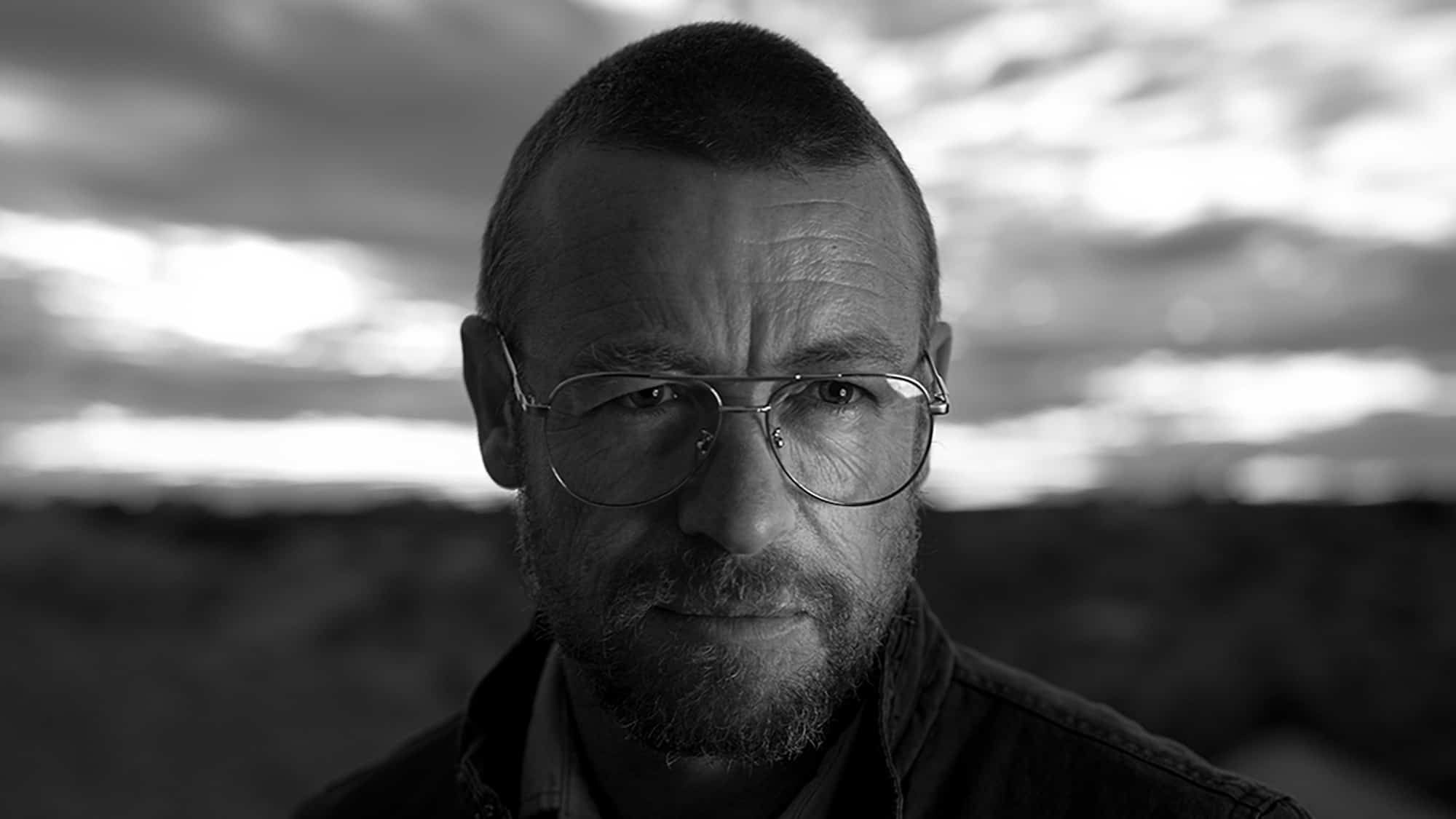 Man with glasses staring intently at the camera against a cloudy sky backdrop.