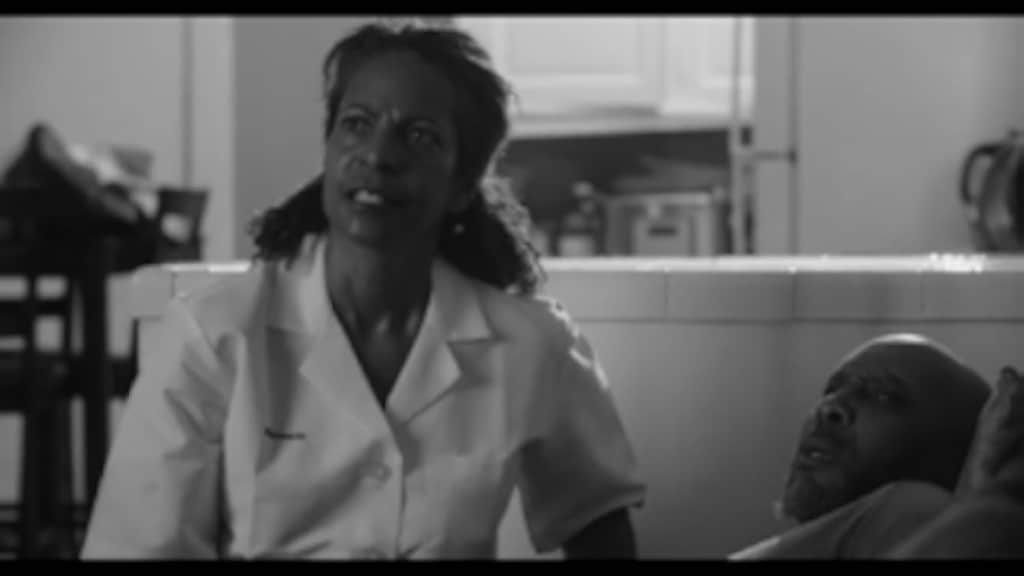 A nurse in a white uniform is standing next to a bedridden patient, expressing concern. the image is in black and white.
