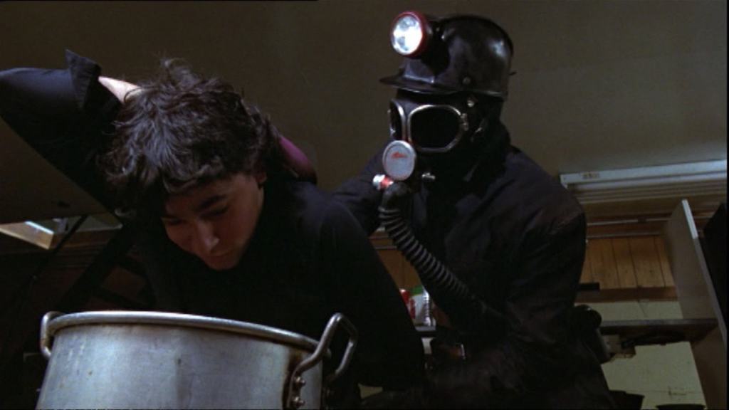 A person in a gas mask and headlamp examines another person's head with a medical device.