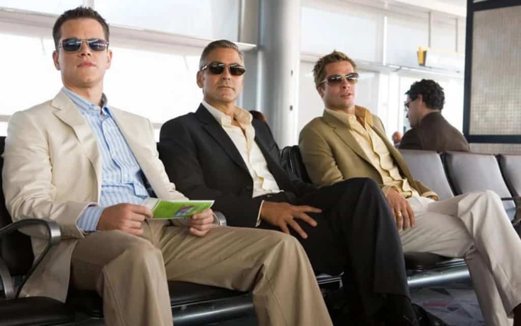 Three men in suits and sunglasses sitting in an airport waiting area.