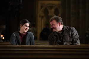 Two people smiling and sitting across from each other on a wooden bench, engaged in a pleasant conversation in a location with ornate interior architecture.