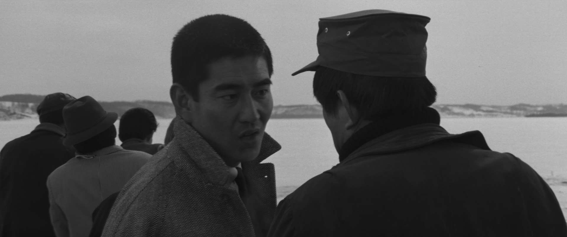 Two men in conversation, with one wearing a military-style cap, captured in a black and white photograph.