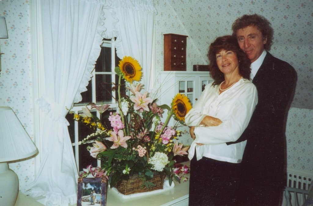 A couple embracing in a room with a bouquet of sunflowers and lilies on the table in front of them.