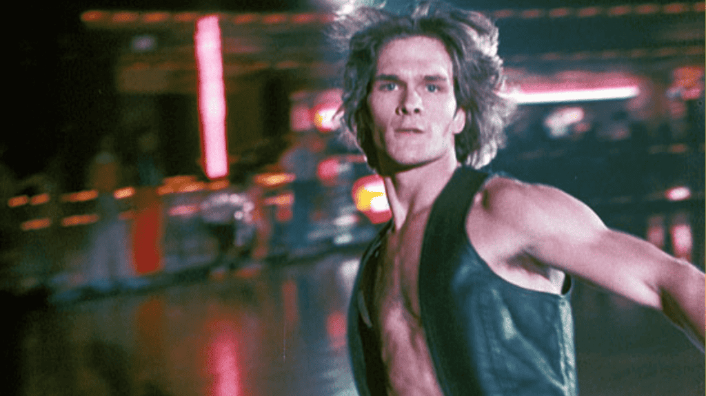 A man with long hair and a sleeveless top appears in motion against a blurred background featuring neon lights.