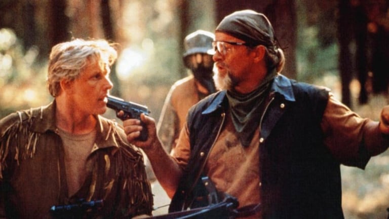 Two individuals in outdoor attire are engaged in a tense standoff in a forest, one pointing a handgun at the other's mouth.