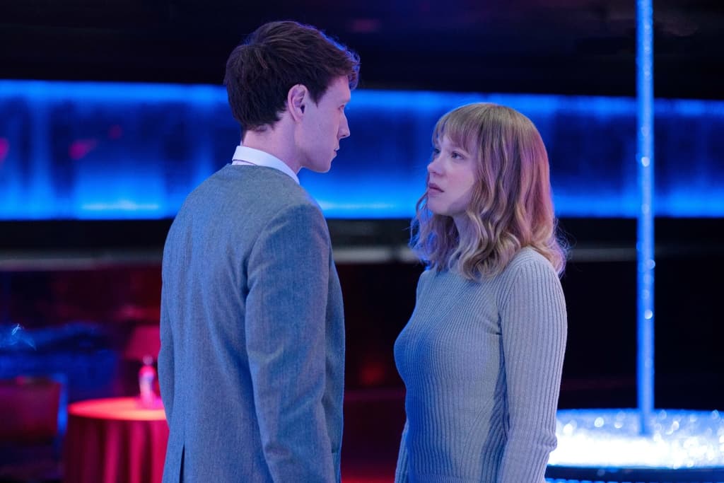 Two individuals facing each other in a dimly lit room with a blue-lit bar in the background.