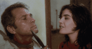 A woman holding a razor to a man's throat in a tense scene.