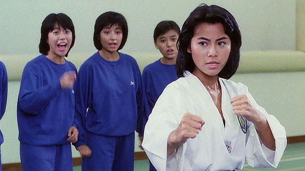 A woman in a karate gi assumes a fighting stance while others in blue uniforms watch with varying expressions.