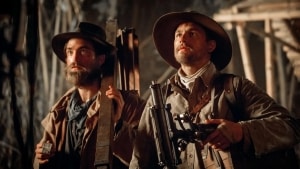Two men in period clothing holding rifles stand alert in a dimly lit wooden structure.
