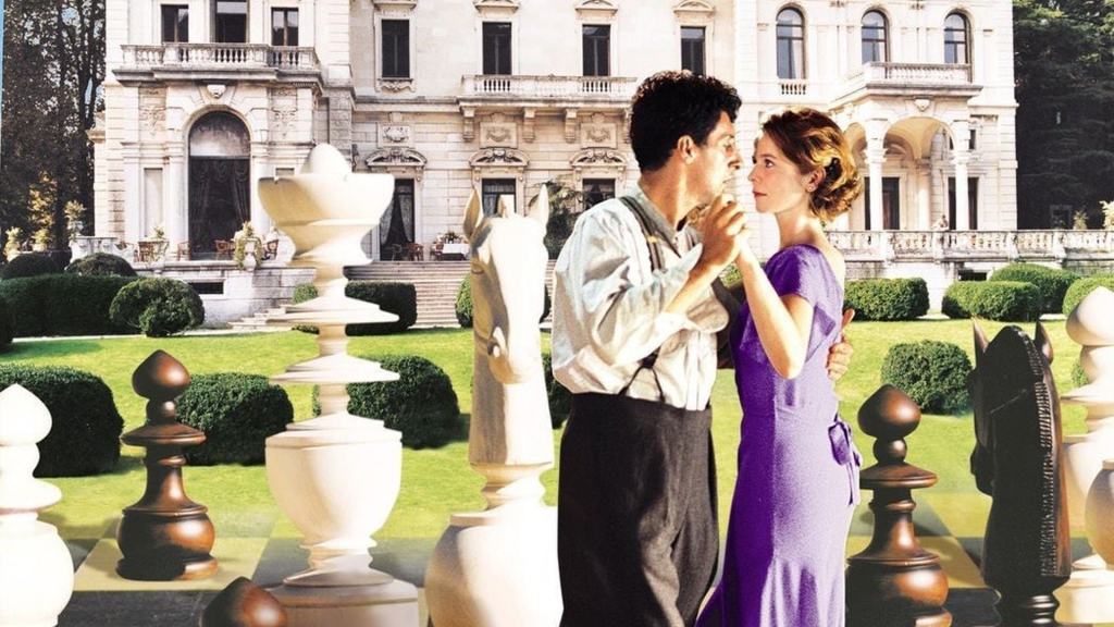 A man and woman standing close together on a giant chessboard in front of an elegant mansion.