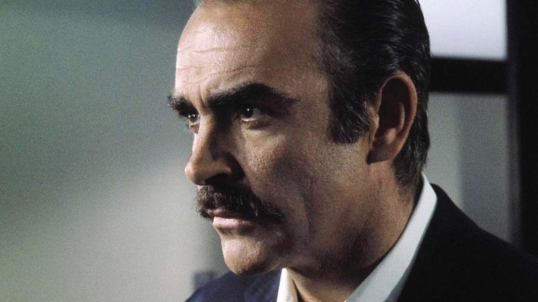 A close-up of a middle-aged man with a prominent mustache, wearing a suit, looking to his left with a serious expression.