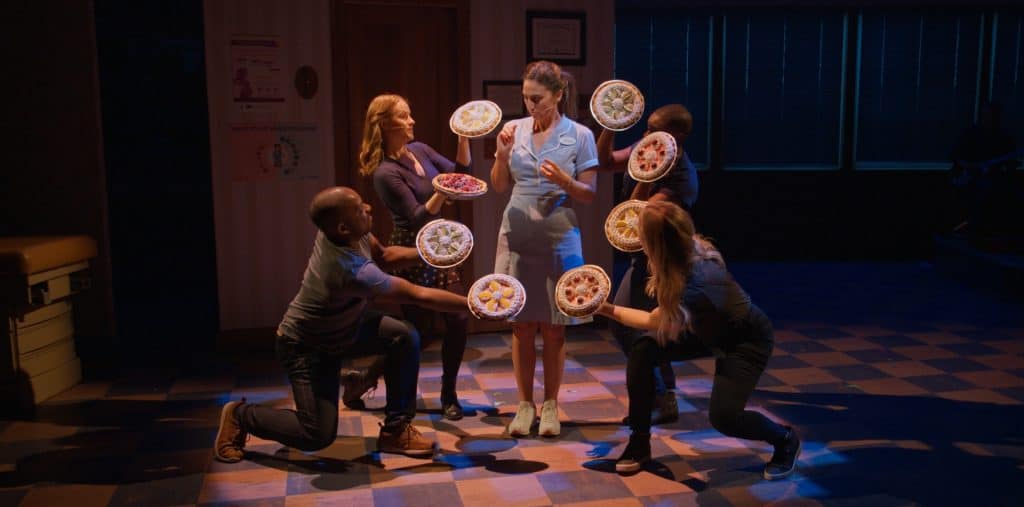 A server in a blue uniform stands with a tray holding several dishes, while three individuals in casual clothing appear to offer her more dishes, in a stylized and theatrical setting.