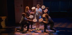 ‘Waitress: The Musical’ Blu-Ray Review – A Transcendent Look At The Heartwarming Broadway Favorite A server in a blue uniform stands with a tray holding several dishes, while three individuals in casual clothing appear to offer her more dishes, in a stylized and theatrical setting.