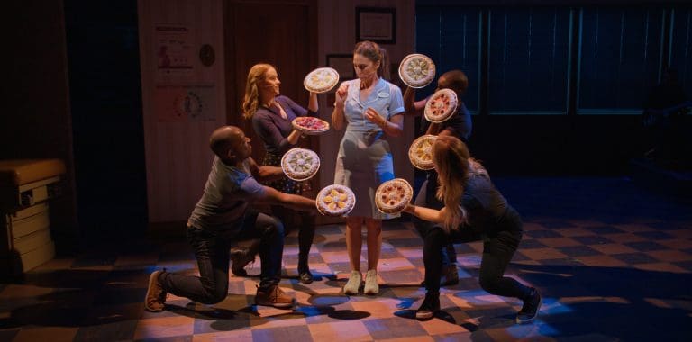 A server in a blue uniform stands with a tray holding several dishes, while three individuals in casual clothing appear to offer her more dishes, in a stylized and theatrical setting.