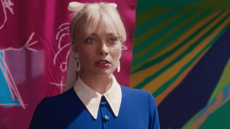 Barbie 2 Is In Trouble Despite Warner Bros. Offering “Life-Changing Money” A woman with blonde hair, wearing a blue dress with a white collar and large earrings, stands in front of colorful, abstract artwork.