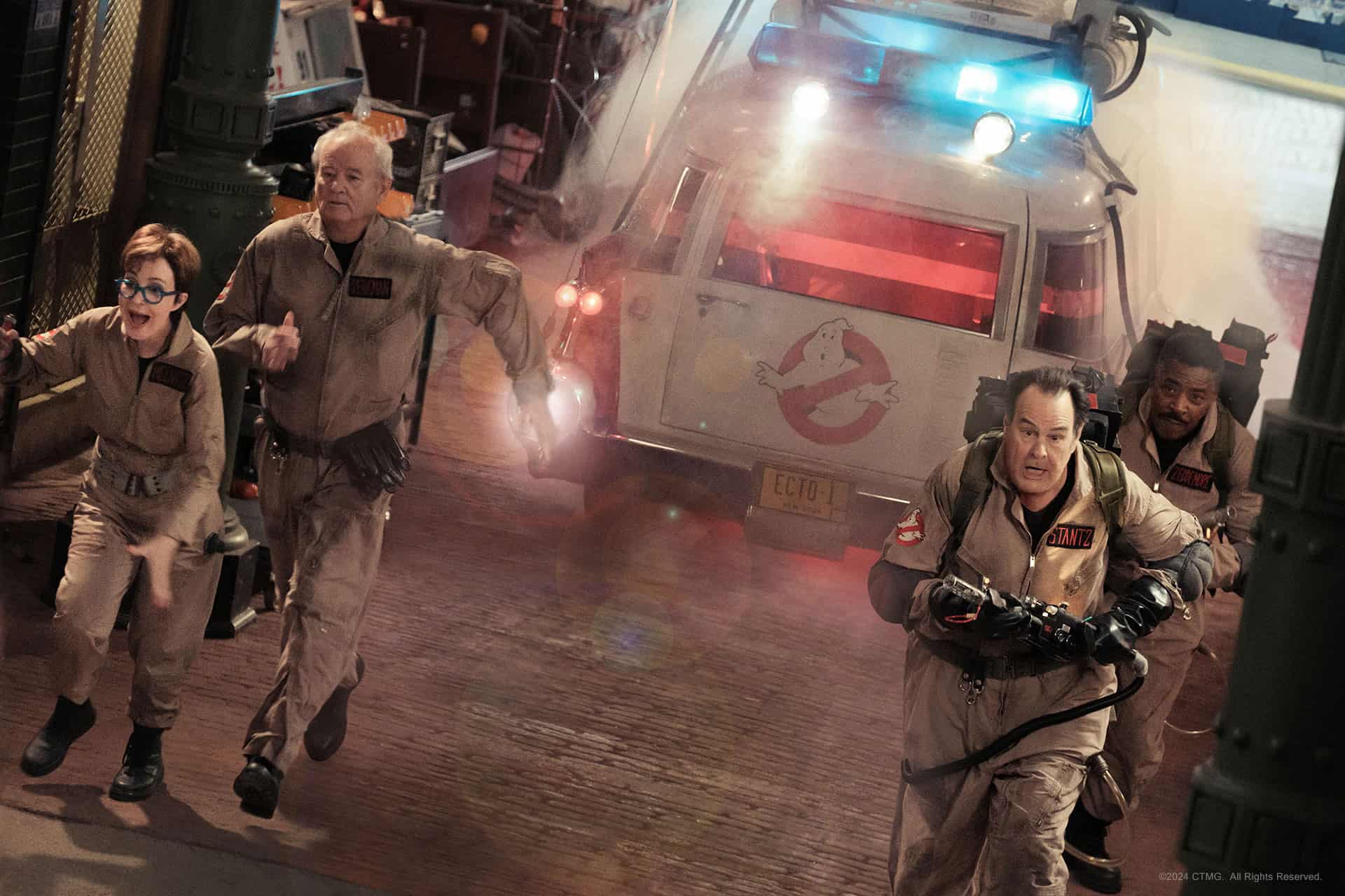 Four ghostbusters in action with their equipment, near their iconic vehicle.