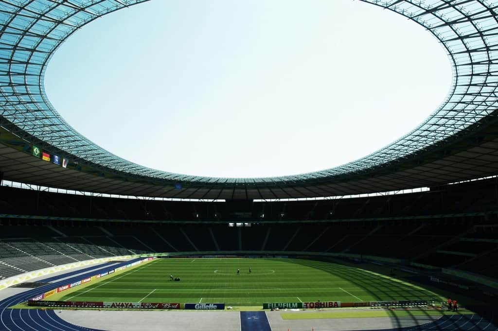 An empty stadium with a large, open roof and a well-maintained grass pitch under a clear sky.