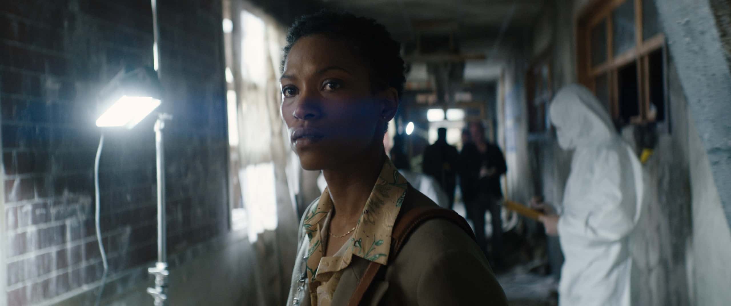 A woman standing in a dimly lit corridor looking to the side with intent, with people in protective gear in the background.