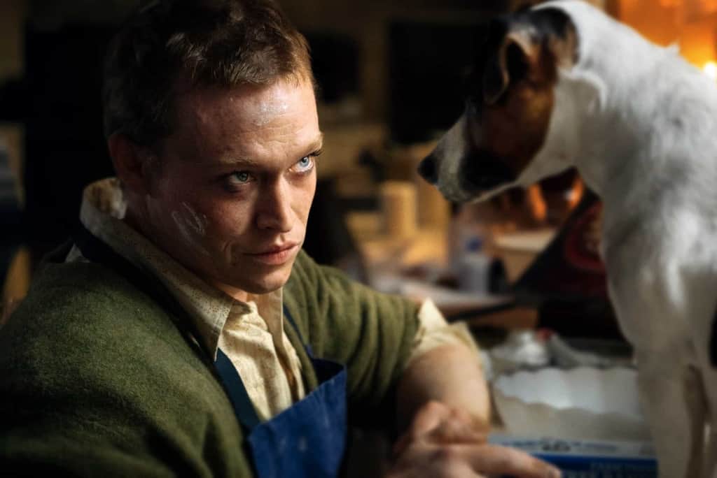 A man in a work apron makes eye contact with a dog standing on a nearby surface.