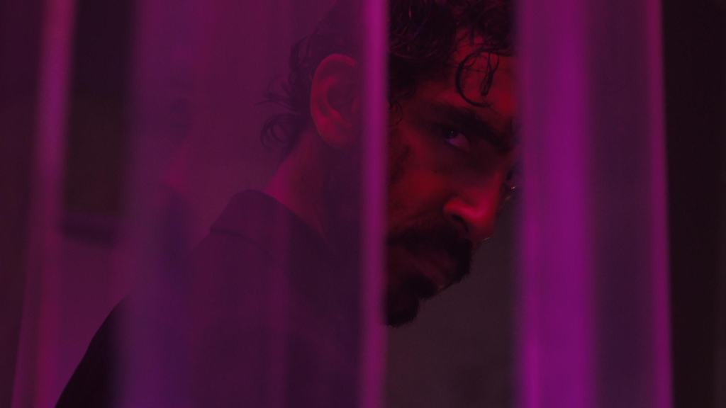 Man with a beard looking intently through purple-tinted bars.