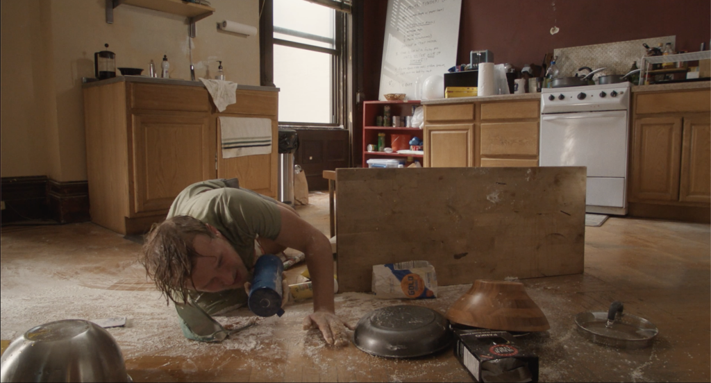 A man lying on a kitchen floor looks under a wooden board, amidst scattered utensils and food containers.