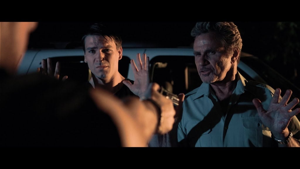 Two men, one younger and one older, stand in front of a vehicle at night with raised hands, facing a person whose hands are partially visible in the foreground.