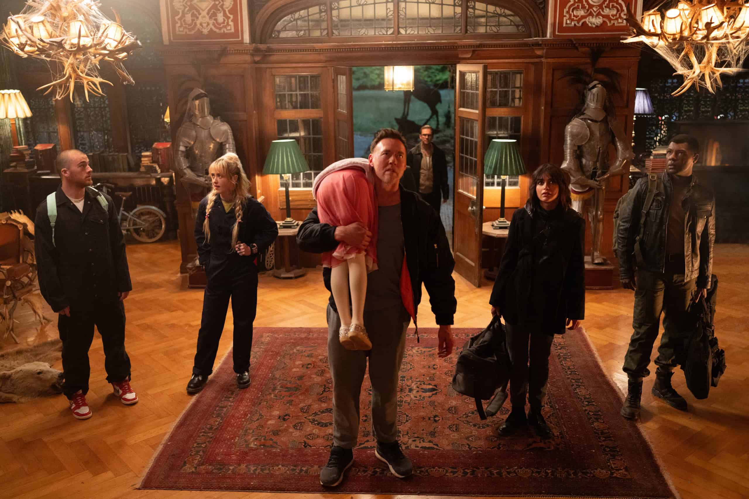 A man carries an unconscious woman in a red jacket inside a grand room, accompanied by four other people, suggesting a tense or urgent situation.