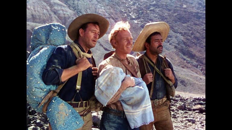 Three men wearing sombreros and backpacks standing in a rocky desert, looking off to the side, as if gazing at something in the distance.
