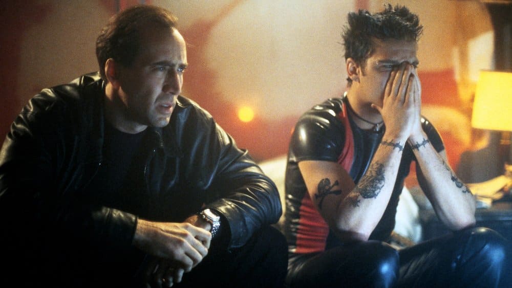 Nicolas cage and james franco sit together in a dimly lit room, with cage appearing concerned and franco covering his face with his hands.