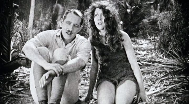 Two individuals in vintage attire sitting on the ground in a wooded area, appearing pensive or in contemplation.