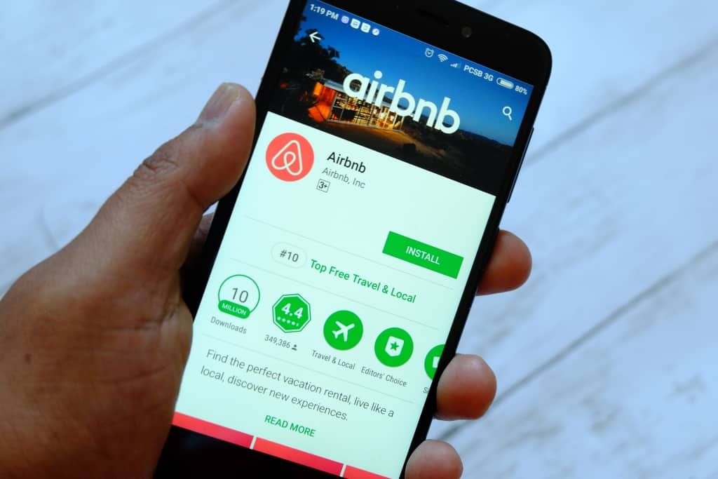 A hand holding a smartphone displaying the airbnb app download page on google play store, with an install button and app details visible.