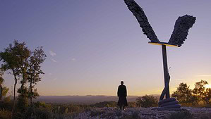 Janus Contemporaries To Release The Wim Wenders Documentary ‘Anselm’ On 3D Blu-Ray This July A man stands beside a large sculpture of a feathered wing at sunset, with panoramic views of a distant mountain range and forest.