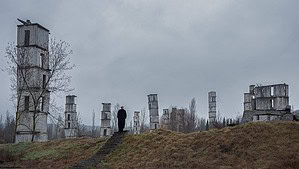A person walks towards dilapidated concrete towers on a grassy hill under a cloudy sky.