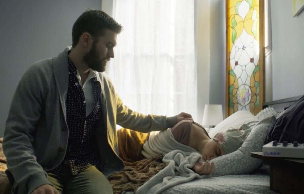 A man seated beside a bed, looking at a young child sleeping under a blanket, in a room with a stained glass window.