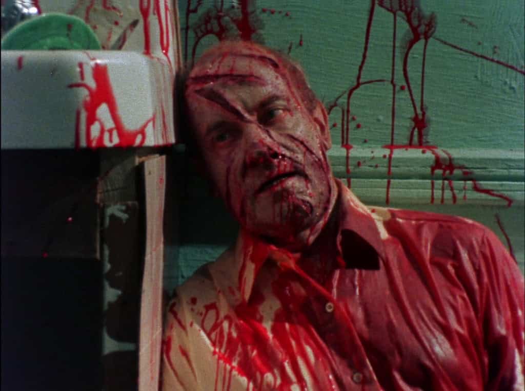 Man with blood on his face and shirt, sitting against a wall with splattered blood, looking distressed.