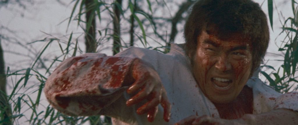 A man with a bloodied face and white shirt, appearing in distress, reaches out urgently through a bamboo thicket.